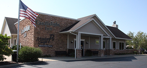Exterior view of Candlelight Inn, a brick and siding building with a gabled roof and columned entrance. A U.S. flag stands in front, with benches, landscaping, and a sign reading "MVP Sports & Grill Company."